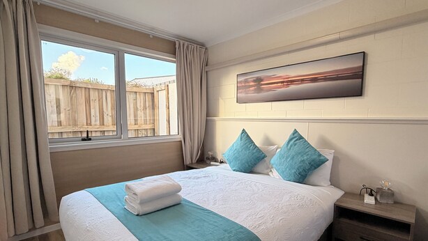 Un dormitorio con una cama grande, una ventana con cortinas, cabecera de madera, una mesita de noche y un cuadro en la pared.
