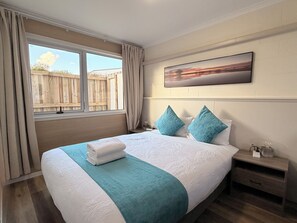 A bedroom with a large bed, a window with curtains, a wooden headboard, a nightstand, and a painting on the wall. - Mountain View Motel, Taupo (Taupo)