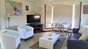 A living room with a TV, white sofas, a coffee table, and a dining area with chairs and a table. - Must Love Dogs B&b (Rutherglen)