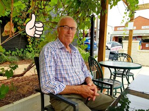 A man sitting at an outdoor table with chairs, surrounded by greenery and a building in the background. - Must Love Dogs B&b (Rutherglen)