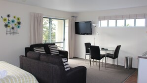 A modern hotel room with a grey sofa, a small dining table with chairs, a TV mounted on the wall, and a window with curtains. - Westport Spa Motel (Westport)