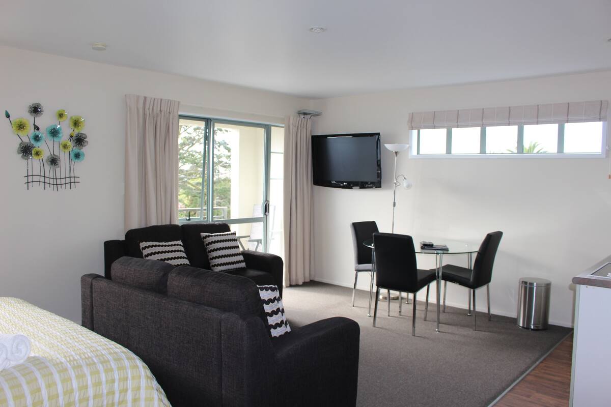 Ein modernes Hotelzimmer mit grauem Sofa, einem kleinen Esstisch mit Stühlen, einem an der Wand befestigten Fernseher und einem Fenster mit Vorhängen.