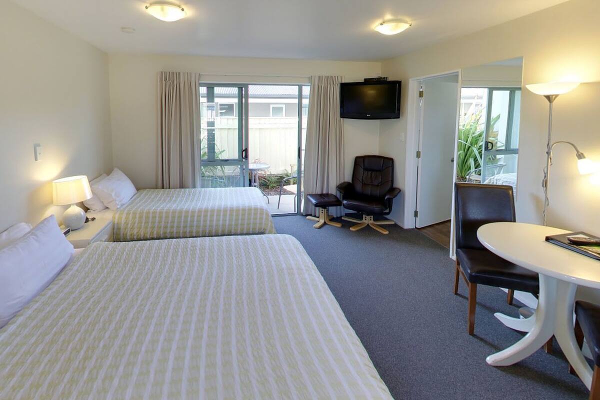 Ein Hotelzimmer mit zwei Betten, einem Fernseher, einem Schreibtisch, einem Stuhl und einem Balkon mit Vorhängen.