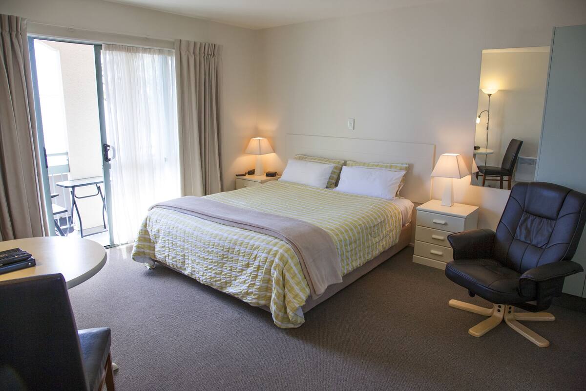 Ein Hotelzimmer mit einem Bett, zwei Nachttischen mit Lampen, einem Schreibtisch mit Stuhl, einem Sessel und einer Schiebetür zum Balkon.