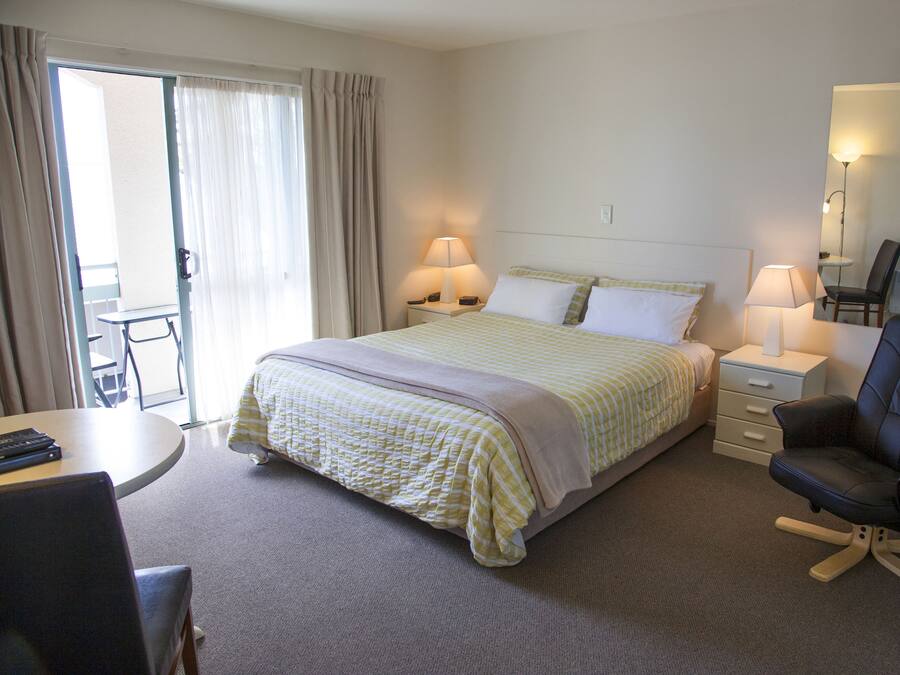 Une chambre d’hôtel comprenant un lit, deux tables de chevet avec des lampes, un bureau avec une chaise, une chaise avec un dossier et une porte coulissante donnant sur un balcon.