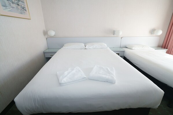 Habitación de hotel con dos camas, cada una con ropa blanca y almohadas, una mesita de noche con lámpara y un cuadro en la pared.