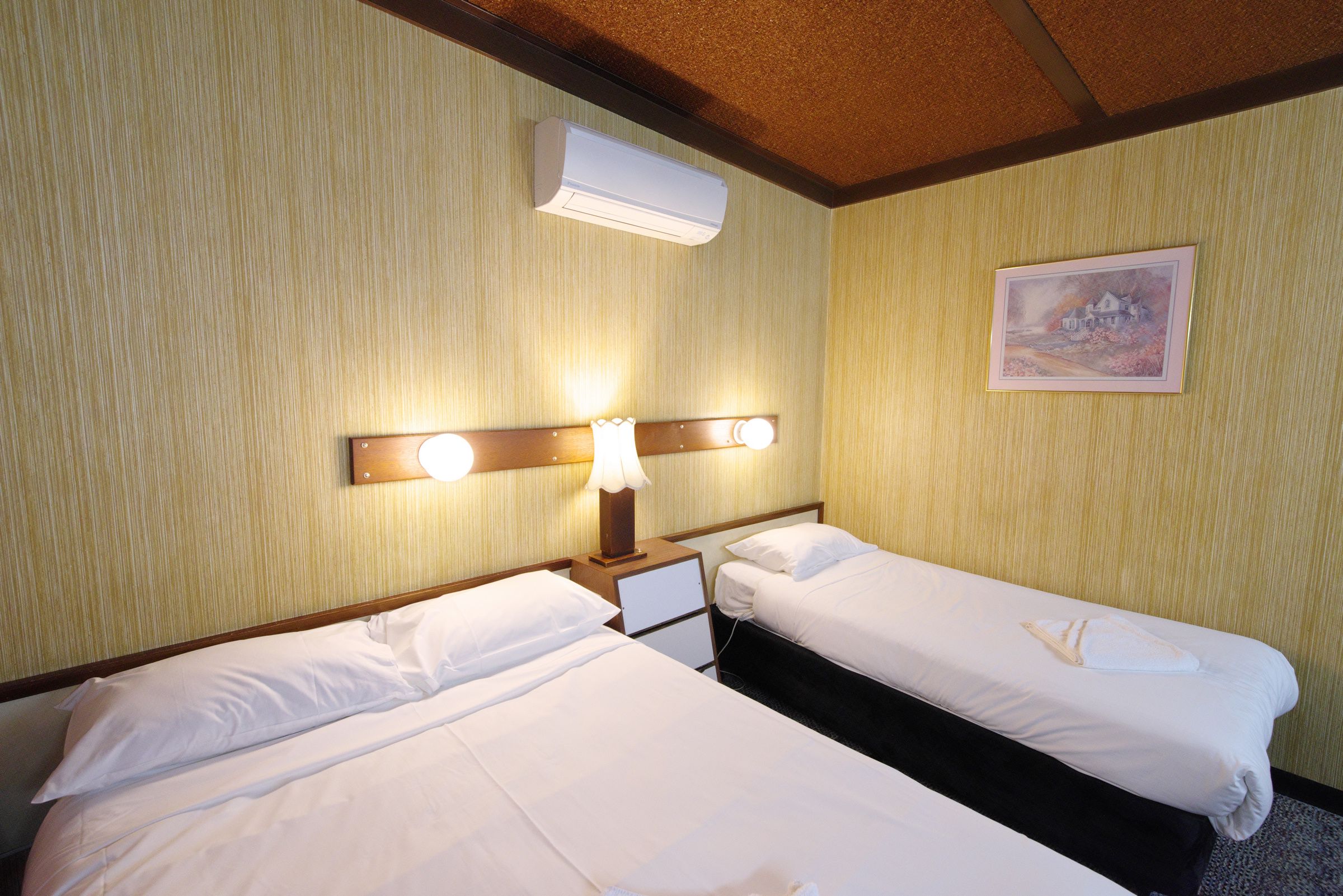 Una camera d'albergo con due letti, un comodino, una lampada, un condizionatore d'aria e un quadro appeso al muro.