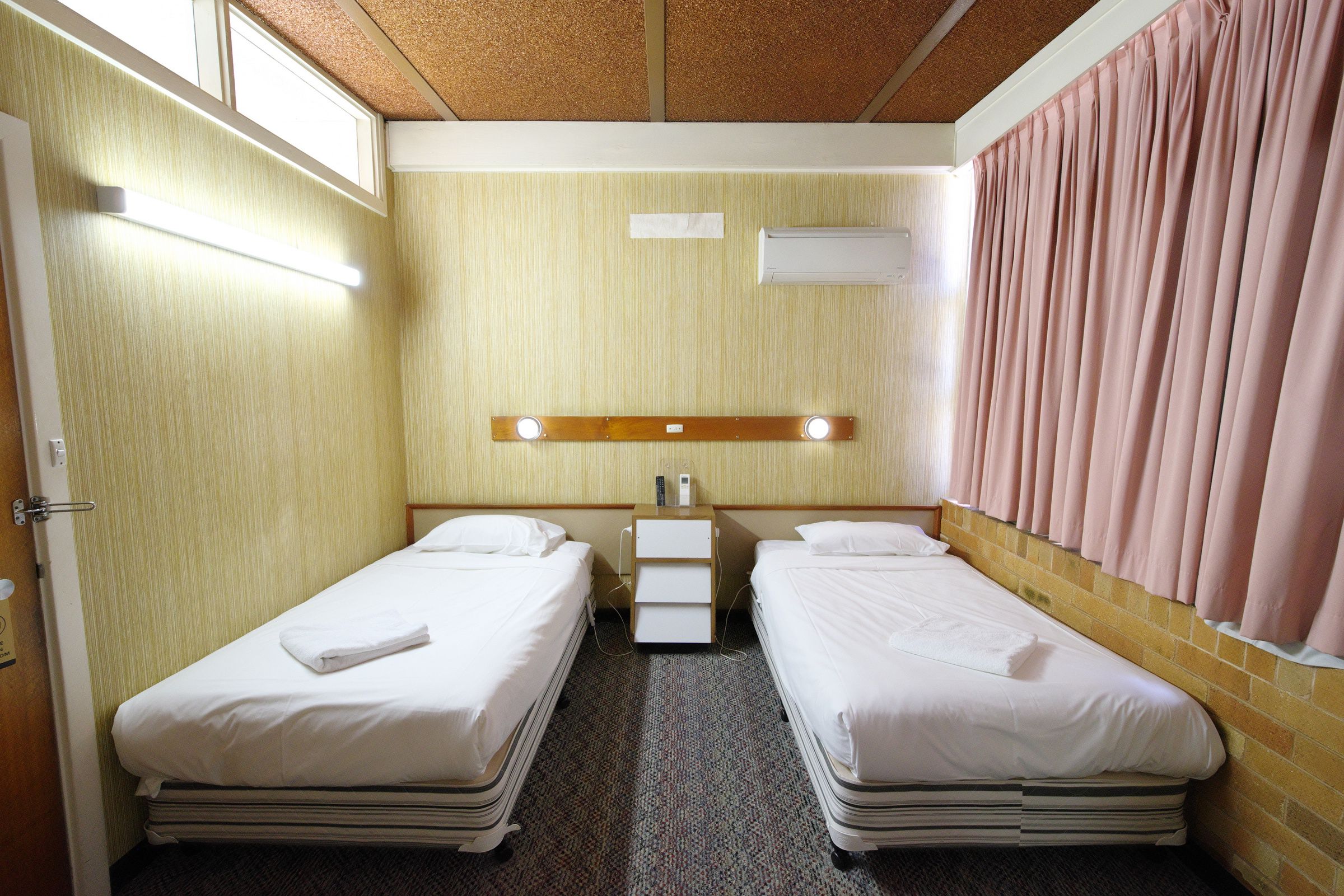A hotel room with two beds, a small bedside table, a wall-mounted air conditioner, and a window with curtains.