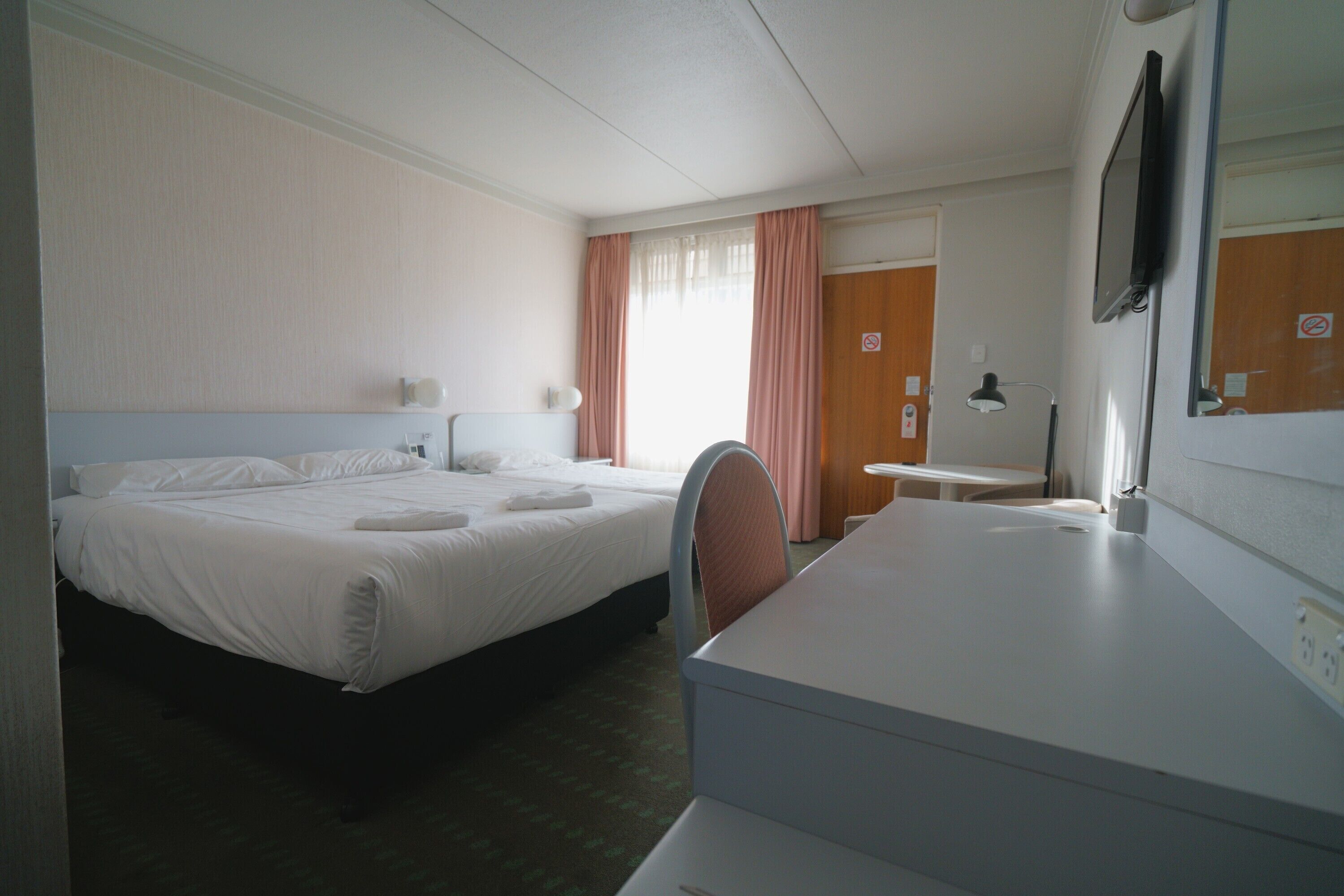 Habitación de hotel con cama, escritorio, silla, televisor y ventana con cortinas.