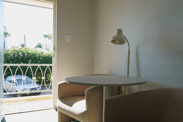A room with a round table, two armchairs, a floor lamp, and a view of a street with cars and greenery outside.