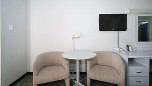 Una habitación pequeña con un escritorio blanco, dos sillas color beige, una mesa blanca, una lámpara, un televisor de pantalla plana y un espejo.