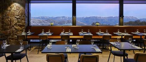 Un restaurante con mesas y sillas dispuestas en filas, amplios ventanales que ofrecen vistas a montañas nevadas.