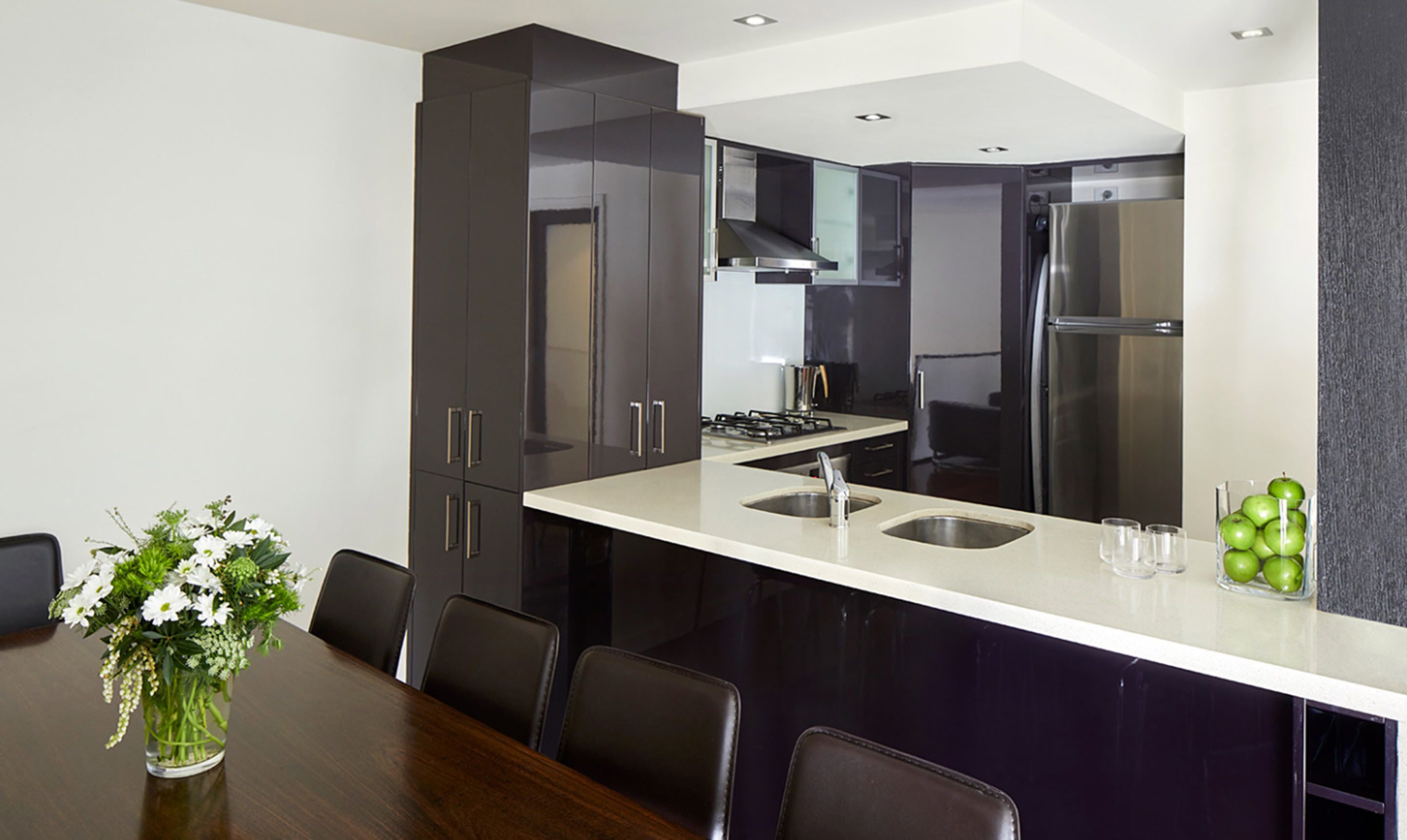 A modern kitchen with dark cabinets, a white countertop, stainless steel appliances, and a dining area with a table and chairs.