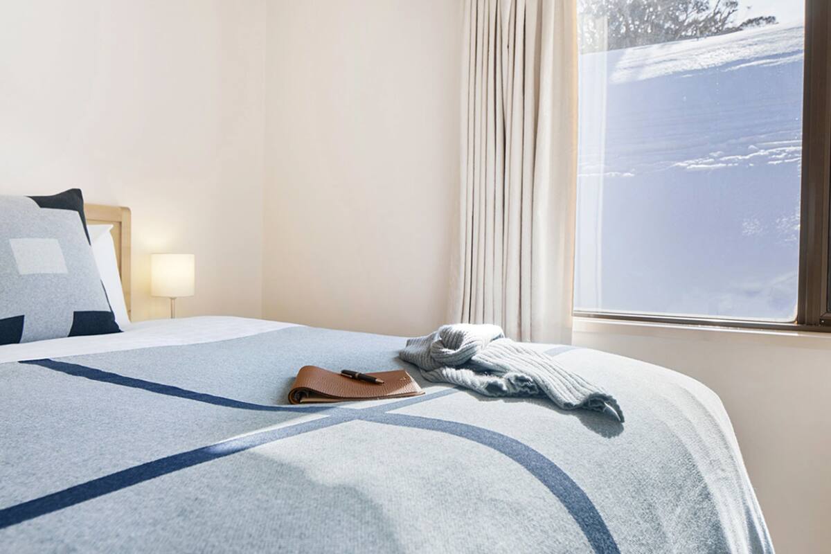A hotel room with a bed, a nightstand, a lamp, and a window with a view of a snowy landscape.