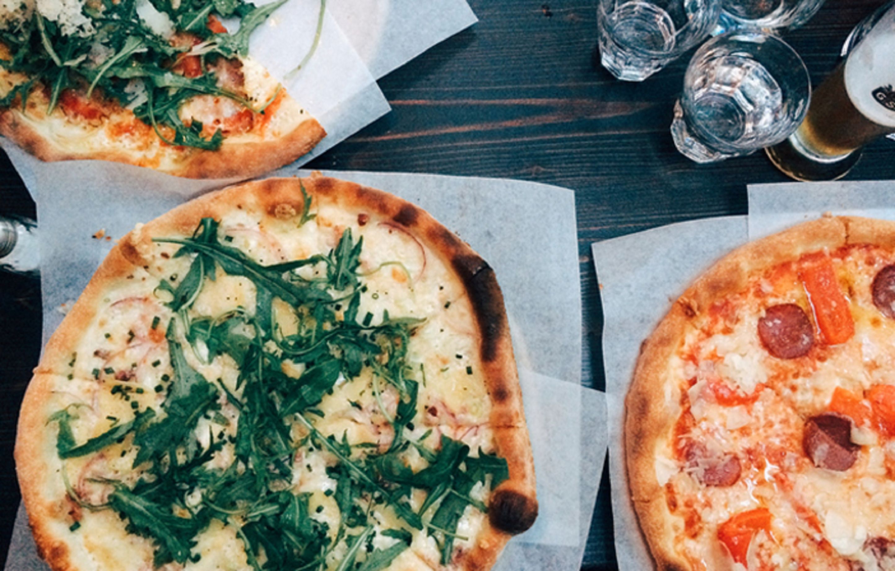 Two pizzas on a wooden table with glasses and a bottle.