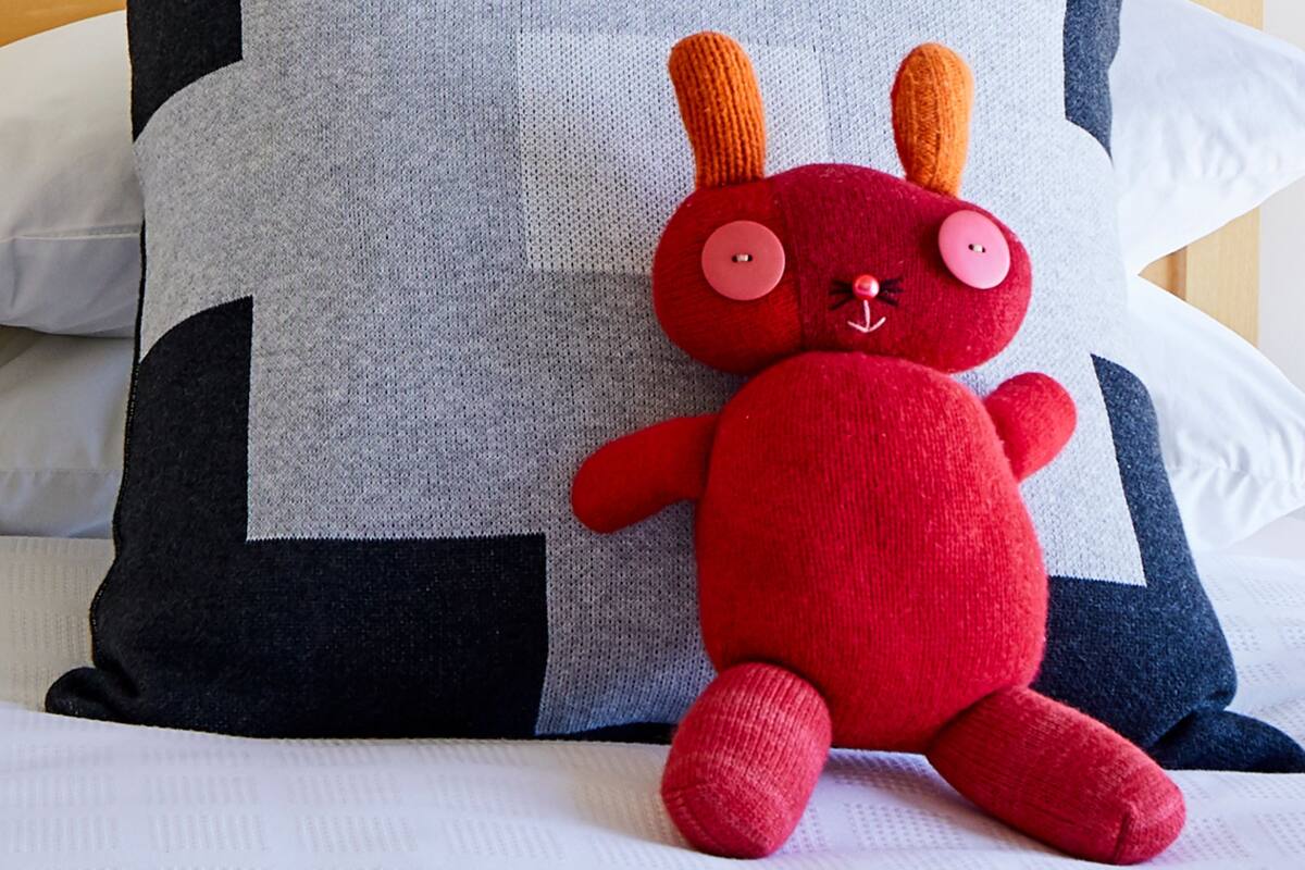 A red plush toy with orange ears sitting on a bed with a grey pillow and a black and white pillow.