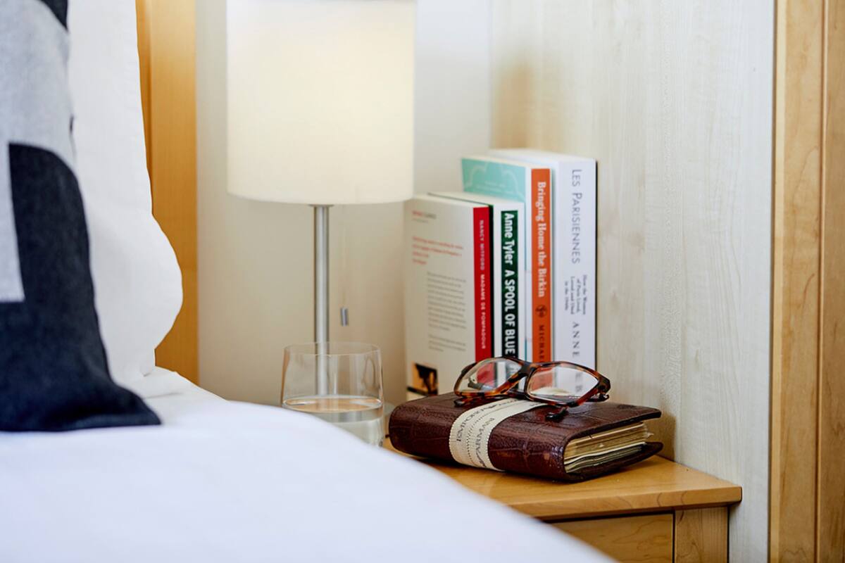 A neatly made bed with a pillow, a bedside table with a lamp, a glass of water, a pair of glasses, and a stack of books.