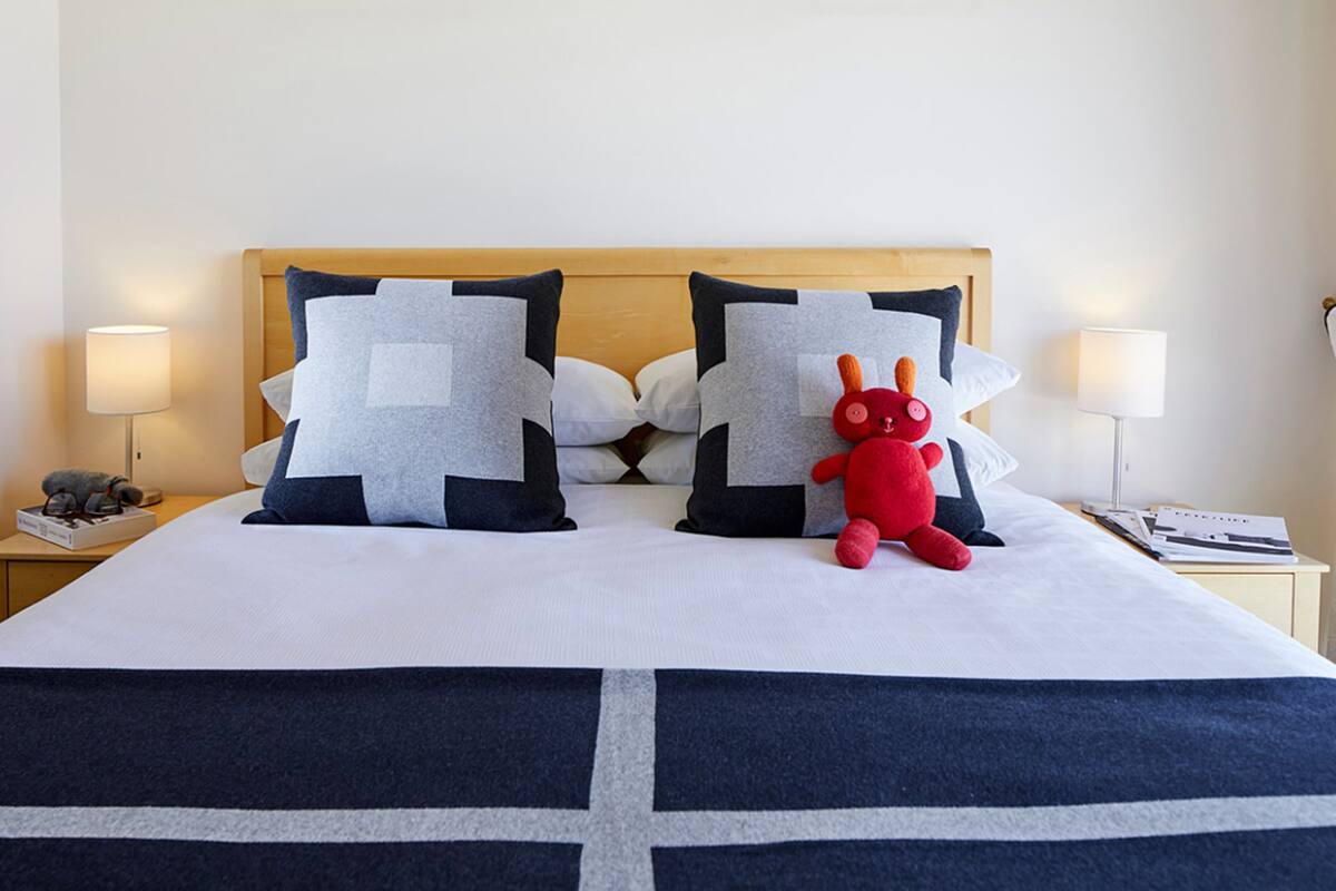 A neatly made bed with a white and navy blue bedspread, two large pillows with geometric patterns, a red plush toy, and two bedside lamps on a wooden nightstand.