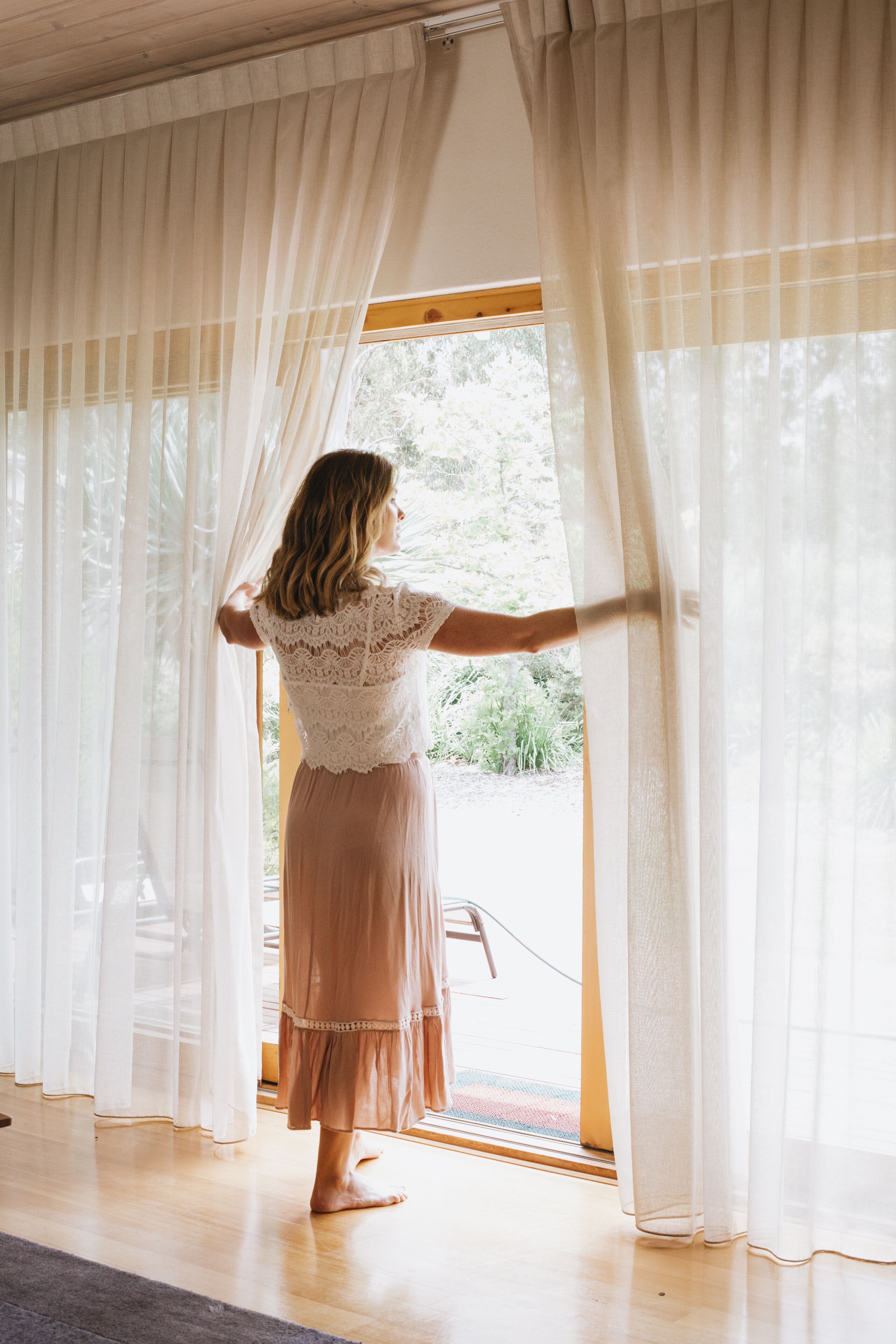 A room with a large window covered by sheer curtains, a wooden floor, and a visible outdoor area with greenery.