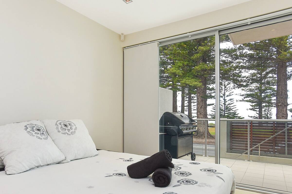 Quarto com uma cama, duas almofadas, um cobertor e vista para árvores através de uma porta de correr de vidro.