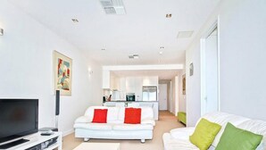 A modern living room with a flat-screen TV, white sofas, and a kitchen in the background. - Breeze Beachfront Apartments (Victor Harbor)