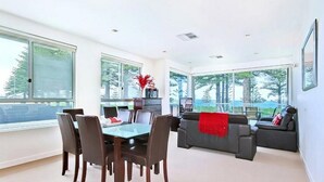 A modern living room with a dining area, large windows, and a view of trees. - Breeze Beachfront Apartments (Victor Harbor)