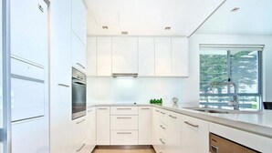 A modern kitchen with white cabinetry, a stainless steel oven, and a window with a view of greenery. - Breeze Beachfront Apartments (Victor Harbor)