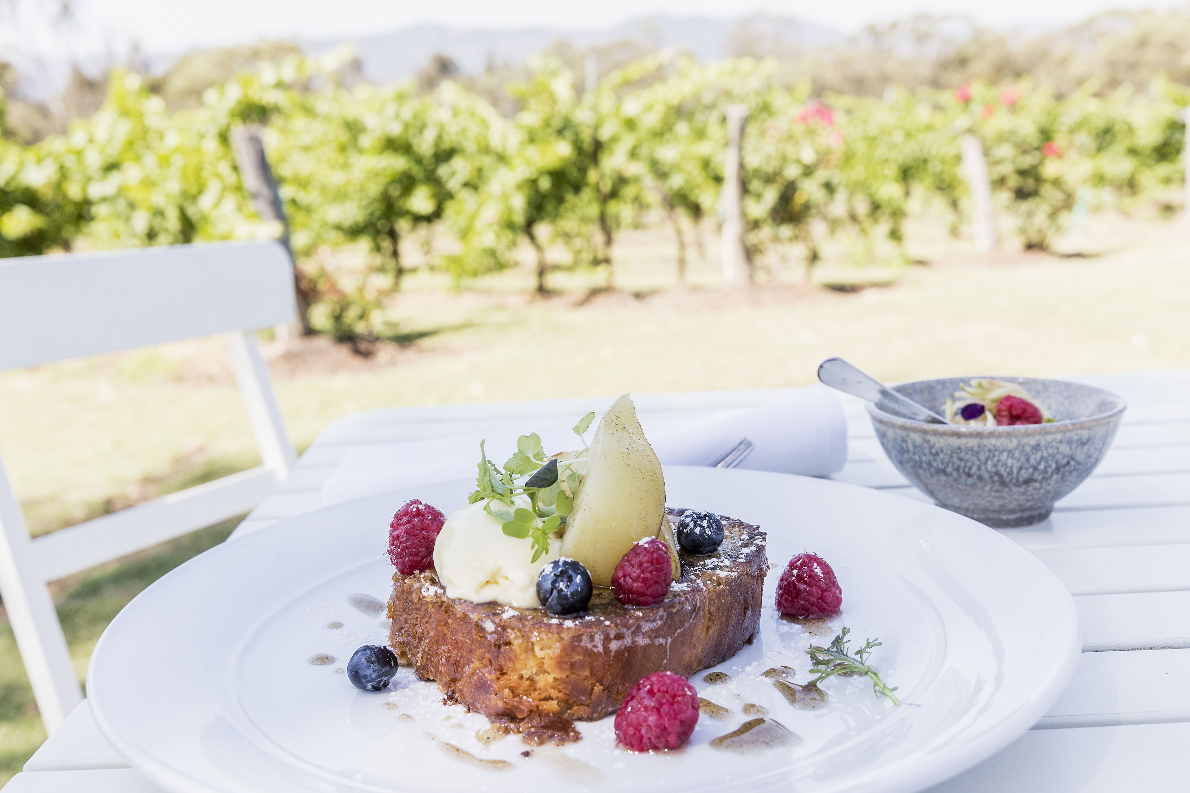 Un dessert servi sur une assiette blanche, agrémenté de fruits rouges et d’une poire, est posé sur une table blanche à l’extérieur, avec un vignoble en arrière-plan.
