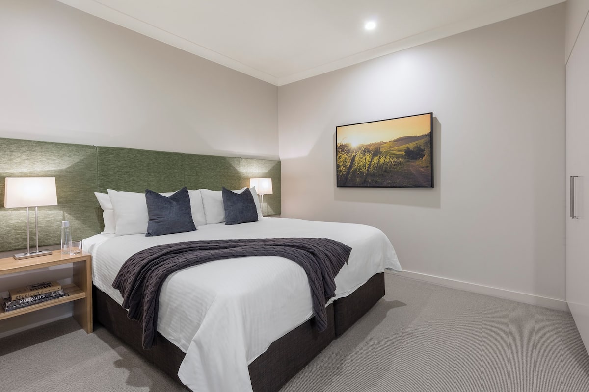 A modern hotel room with a large bed, a green headboard, two bedside lamps, a nightstand with books, and a framed landscape painting on the wall.