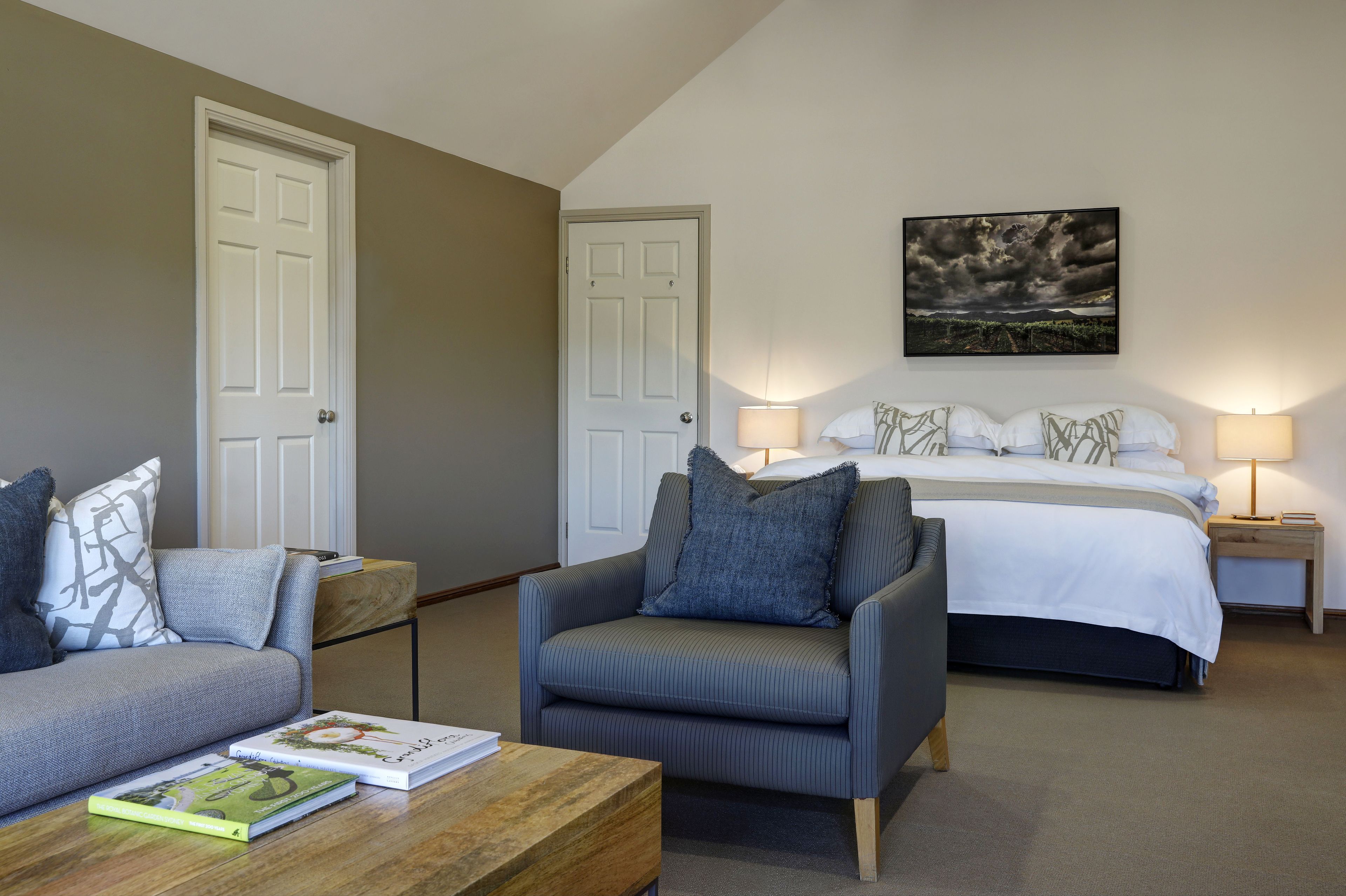 A bedroom with a bed, two bedside tables with lamps, a grey armchair, a wooden coffee table with books, and a painting on the wall.