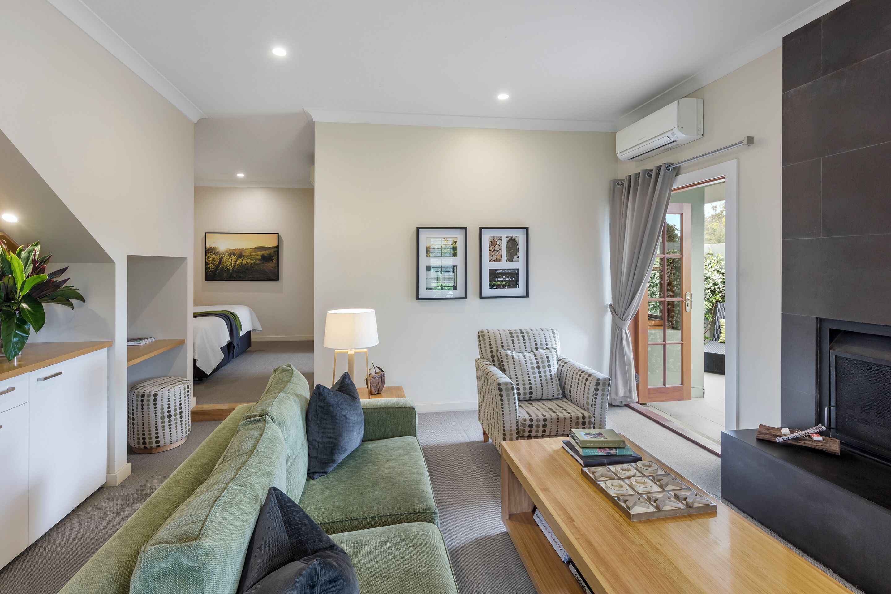 Una sala moderna con un sofá, un sillón y una chimenea. Se observa una cama al fondo y una ventana con cortinas.