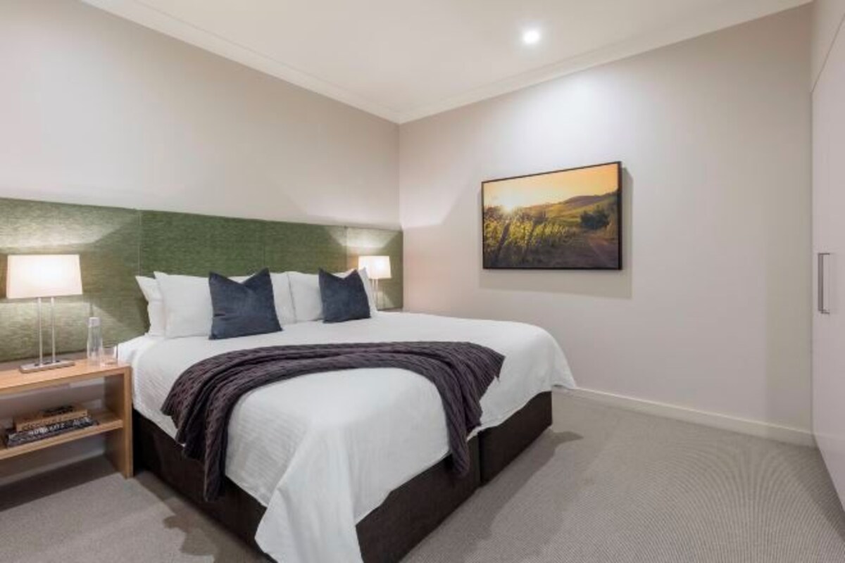 Un dormitorio ordenado con cama, mesitas de noche, cabecera y un cuadro colgado en la pared.