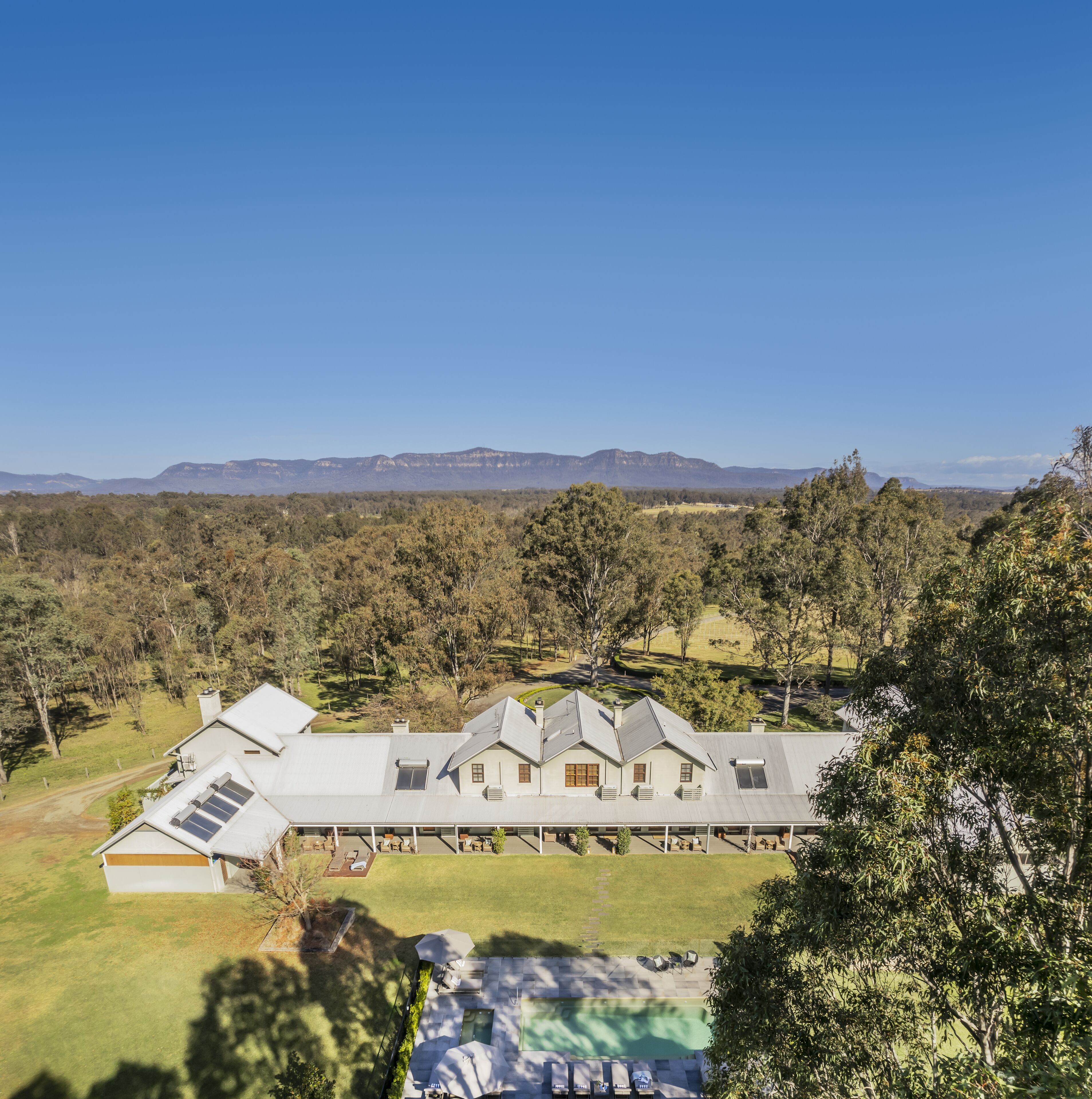 A large, modern house with multiple rooms and a swimming pool, surrounded by lush greenery and mountains in the background.