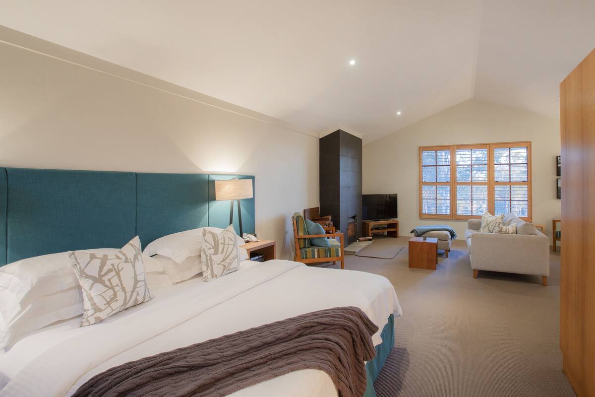 A modern hotel room with a large bed, a seating area with a sofa and armchairs, a television, and a fireplace.