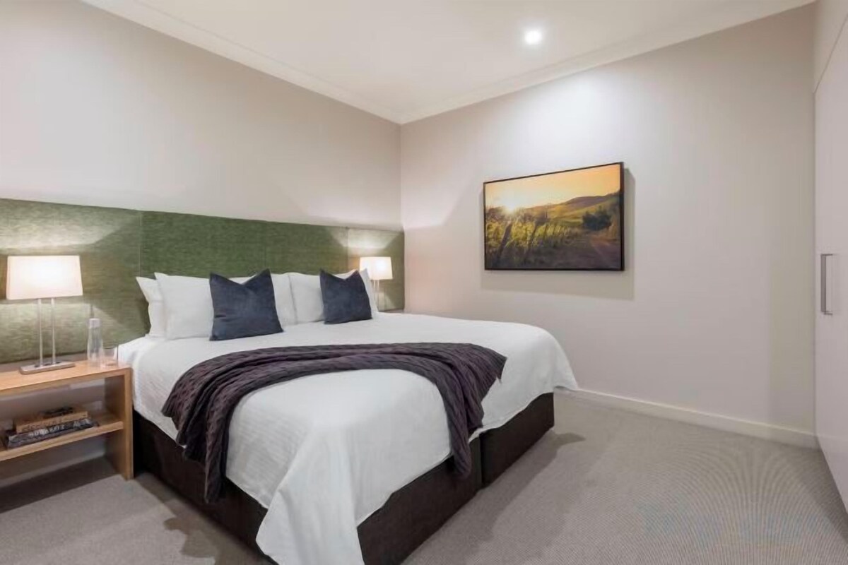 A hotel room with a bed, bedside lamps, a nightstand, a painting on the wall, and a carpeted floor.