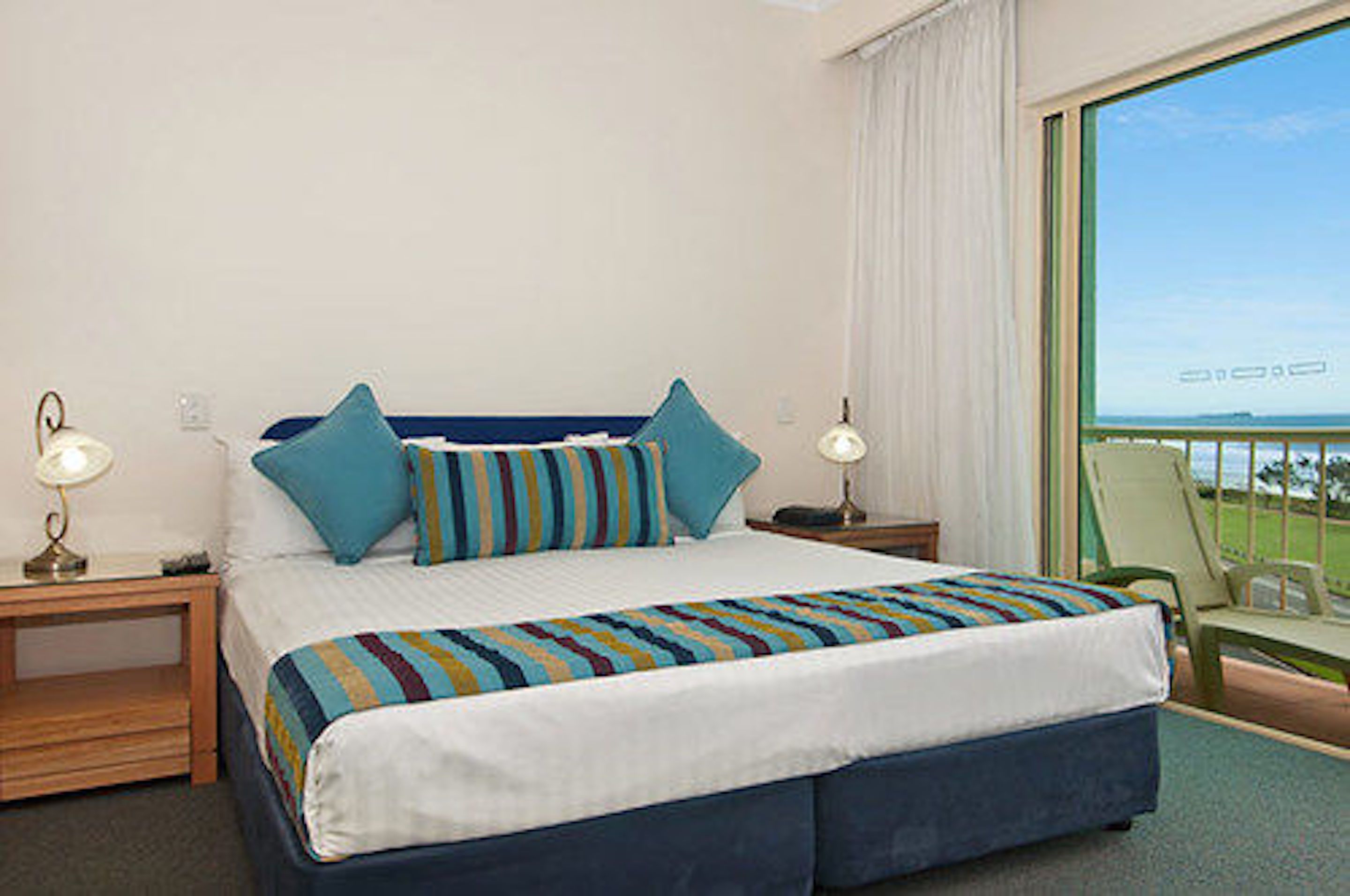 A hotel room with a bed, two bedside tables, a chair, and a view of the sea.