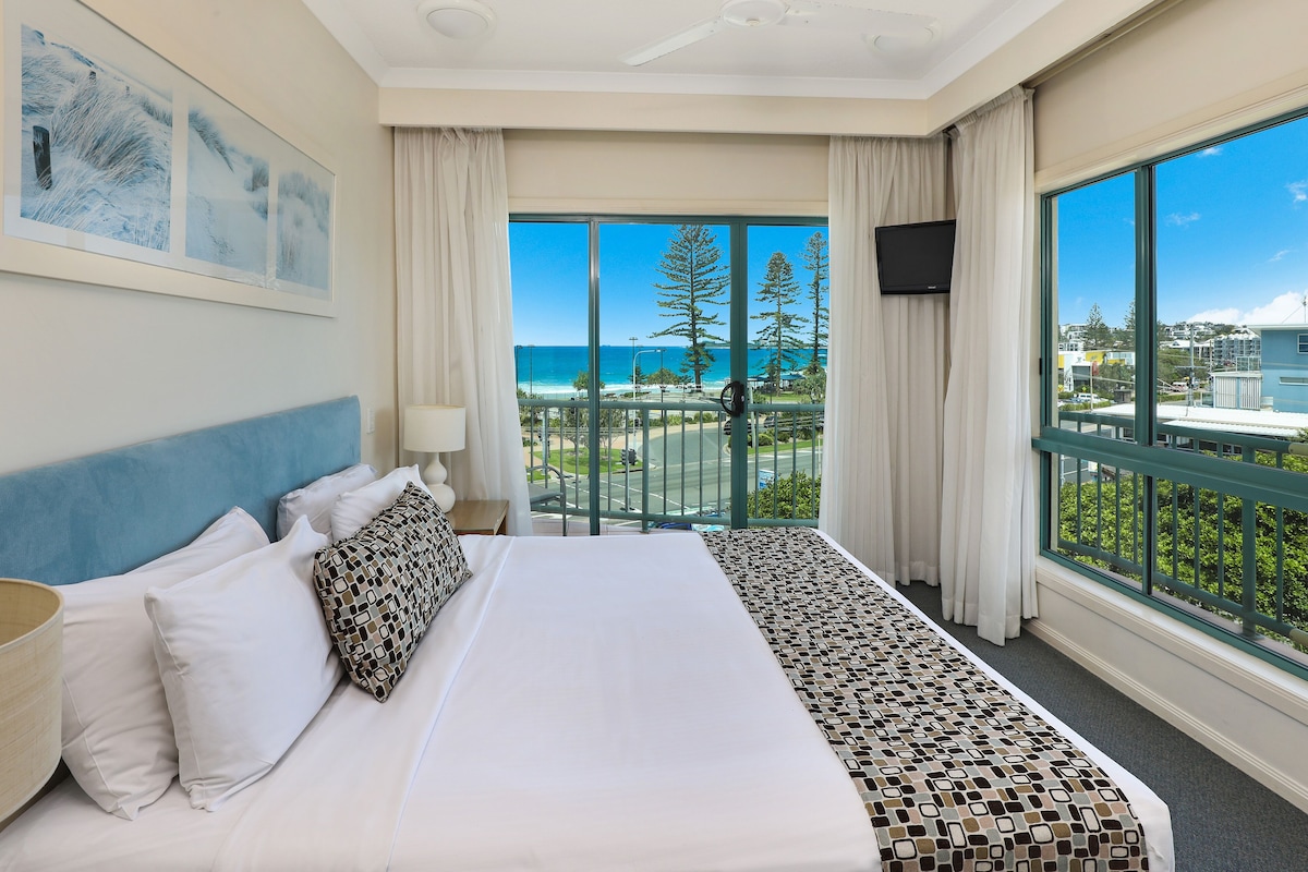 Una habitación de hotel con una cama grande, un televisor y un balcón con vista a la playa y árboles.