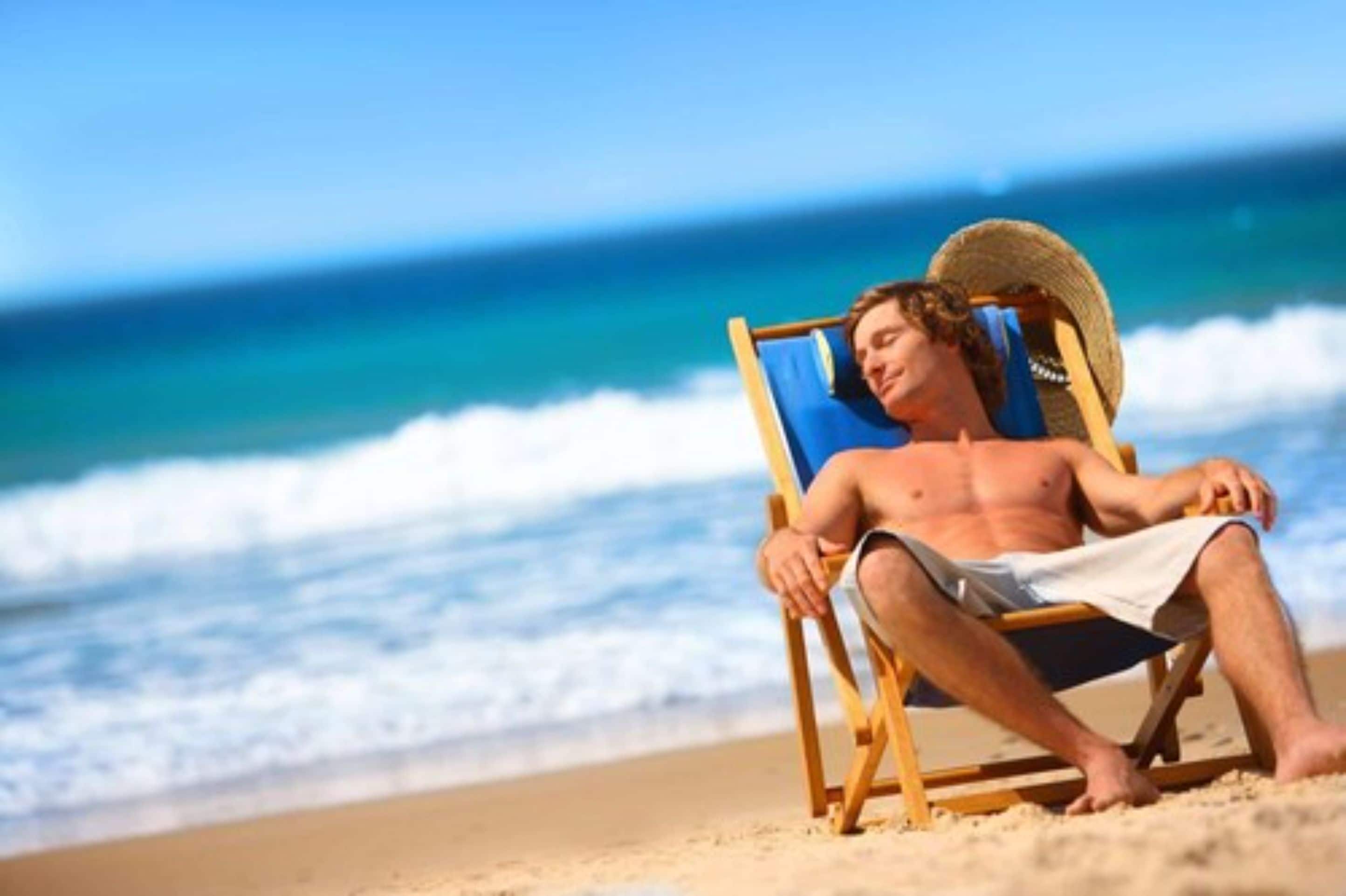 A person sitting on a beach chair with a straw hat, facing the ocean.