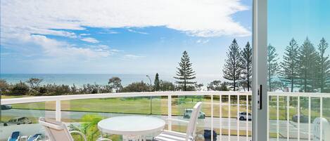 A balcony with a pool, chairs, and a table overlooking a coastal view.