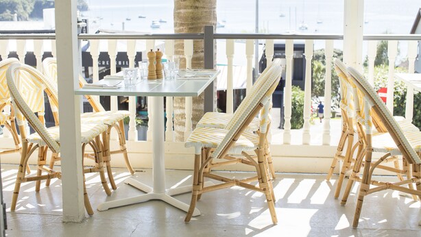 A balcony with wicker chairs and a table, overlooking a coastal view with boats.