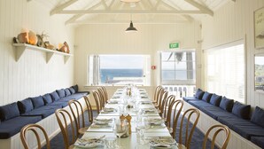 A long dining table with chairs, a high ceiling with wooden beams, and a view of the ocean through large windows.