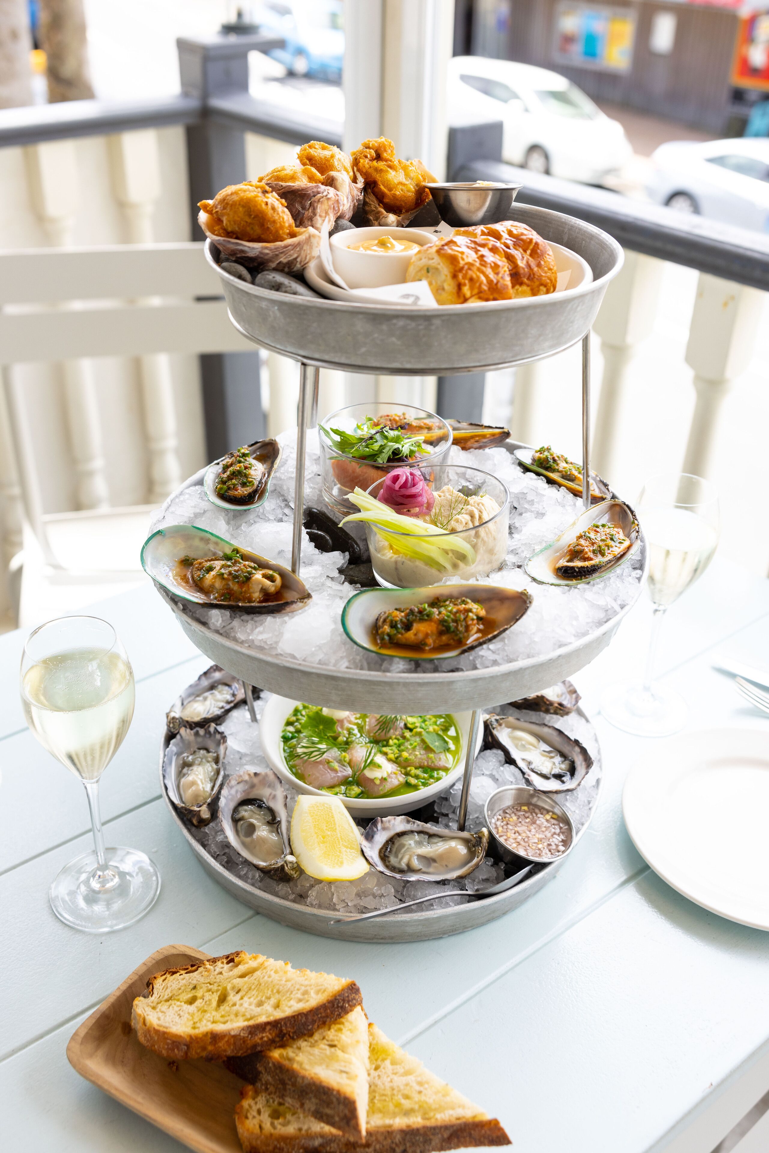 A multi-tiered seafood platter with oysters, bread, and a glass of white wine on a table.