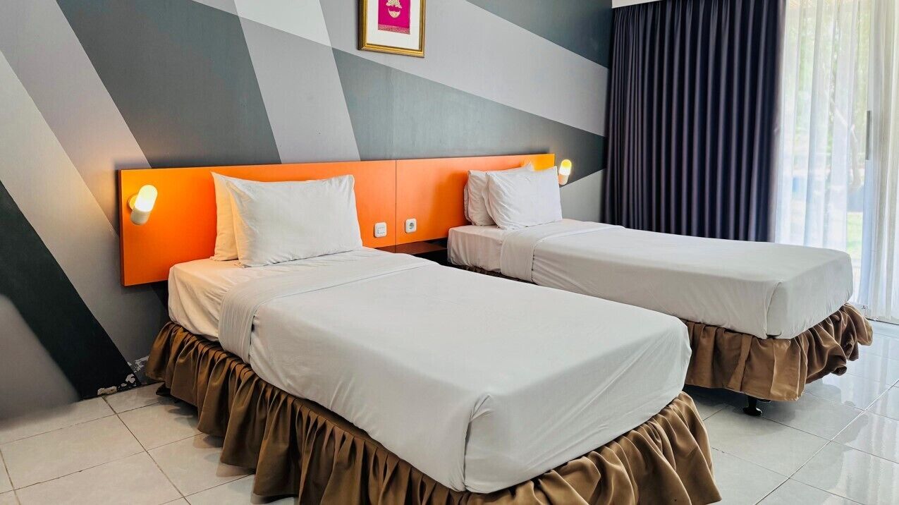 A hotel room with two beds, each with a brown skirted bedspread, a modern orange headboard, and a wall-mounted light.