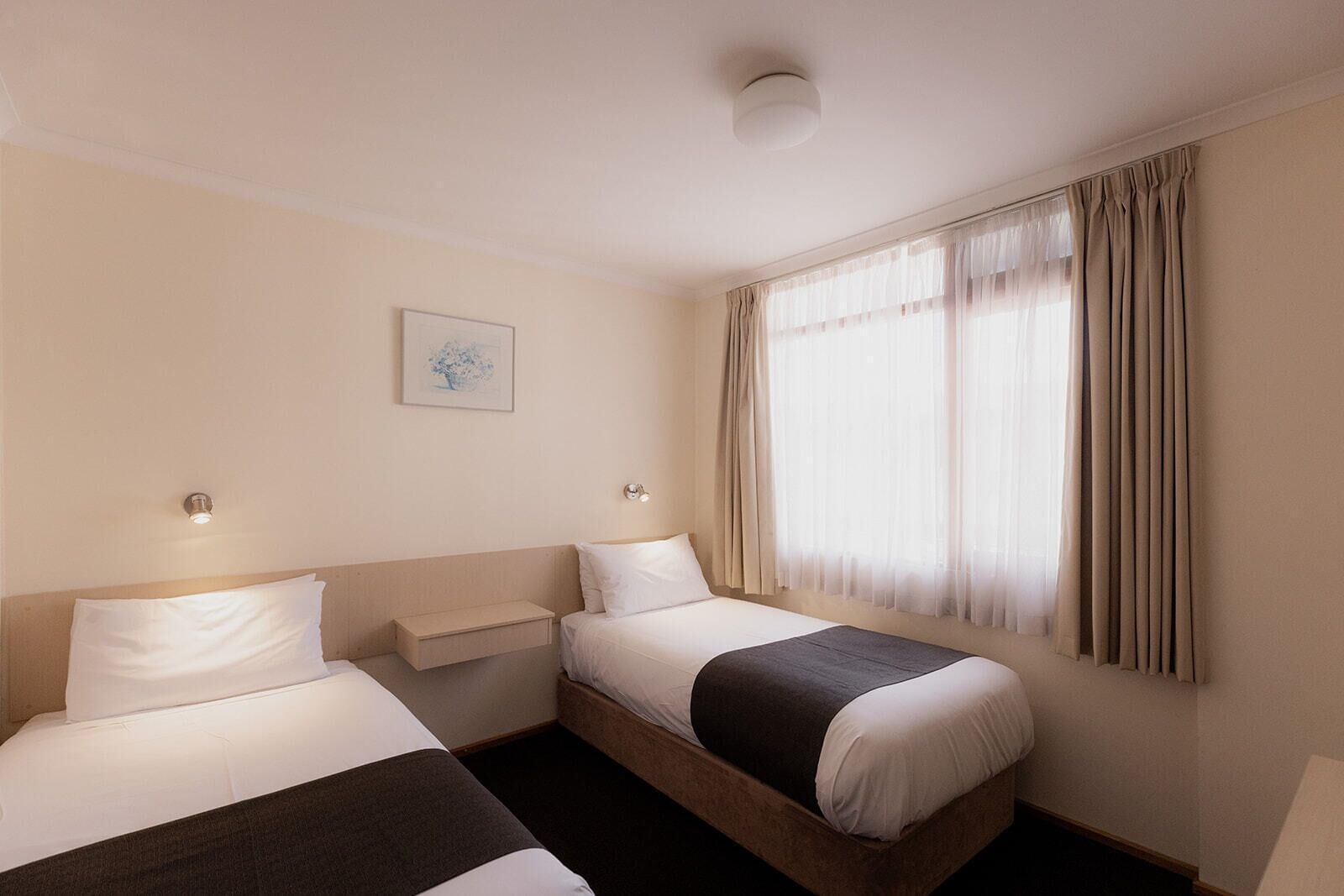 A hotel room with two beds, a bedside table, a small bedside lamp, a window with curtains, and a ceiling light.