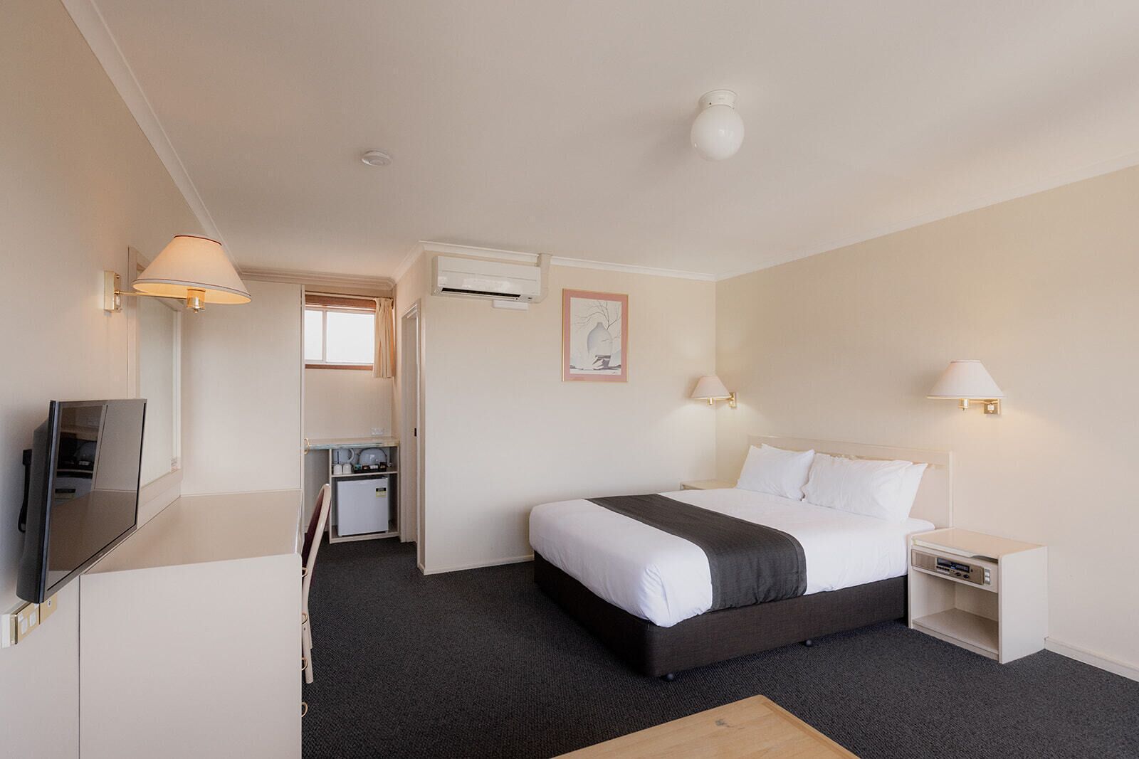A hotel room with a bed, bedside tables, a TV, a desk with a chair, and an air conditioning unit.