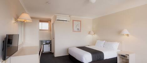A hotel room with a bed, bedside tables, a TV, a desk with a chair, and an air conditioning unit.