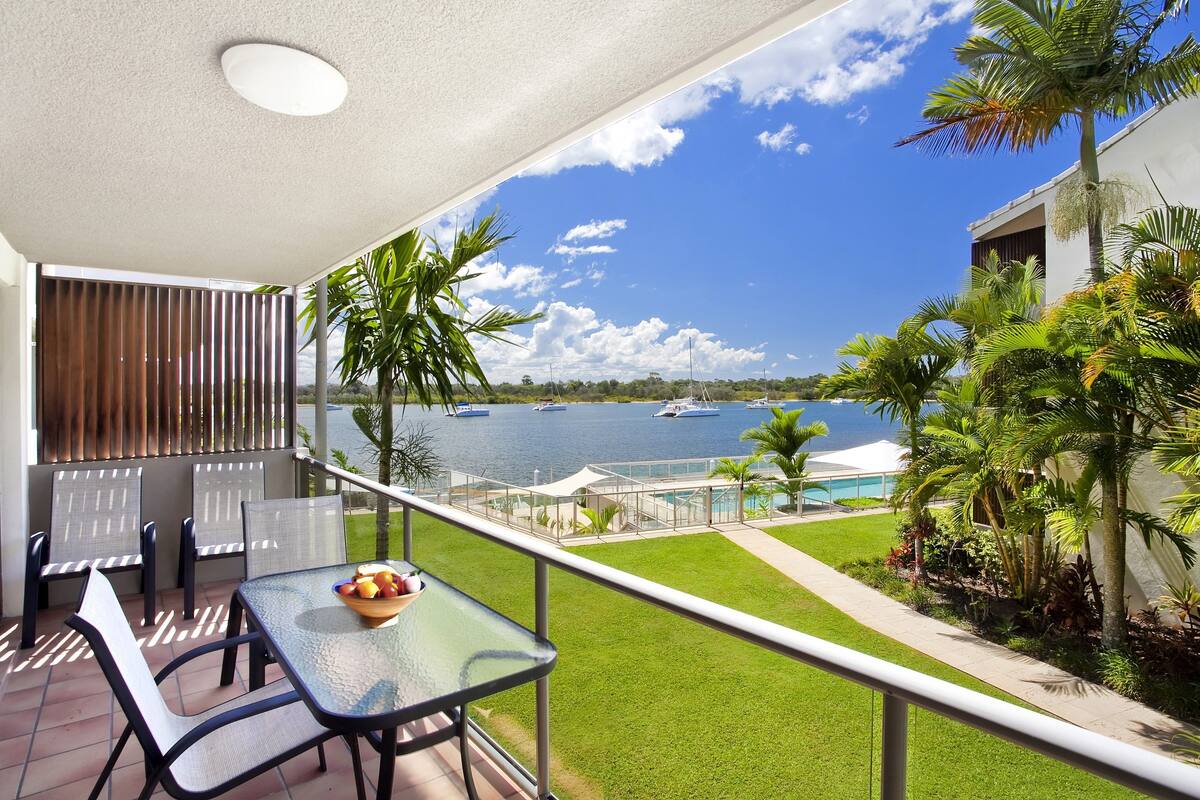 Uma varanda com mesa e cadeiras, vista para o mar com barcos e área da piscina.