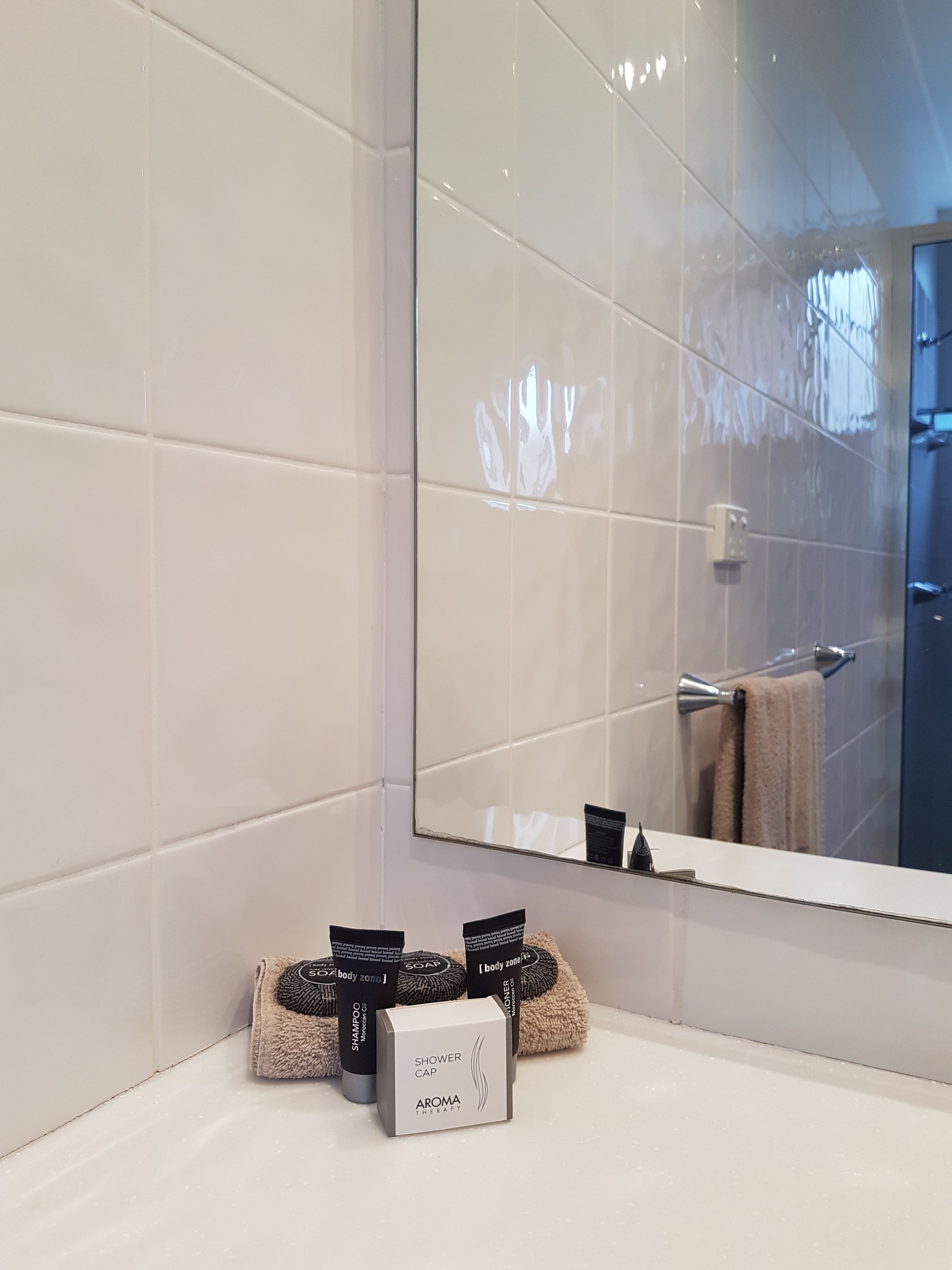A bathroom with white tiles, a large mirror, and a shower cap box labeled "SHOWER CAP AROMA THERAPY" on the counter.