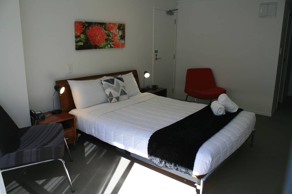 A hotel room with a bed, a chair, a desk, a lamp, and a painting on the wall.
