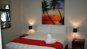 A neatly made bed with a red blanket, two bedside lamps, and a painting of a sunset with palm trees on the wall. - Atlas Suites And Apartments (Tauranga)